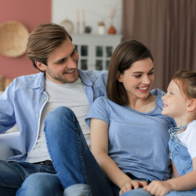 Positive friendly young parents with smiling little daughter sitting on sofa together while relaxing at home on weekend